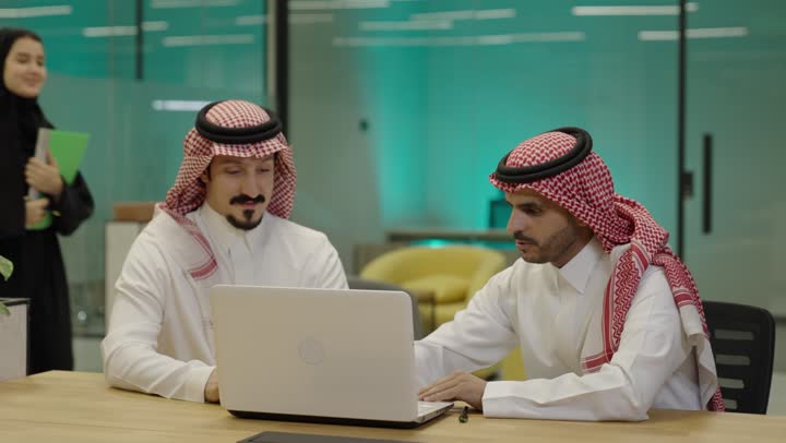 إنجاز الأعمال المطلوبة، دمج الأعمال مع التقنية و التكنولوجيا، رجلان عربيان خليجيان سعوديان يرتديان الزي التقليدي يجلسان في المكتب يعملان على الحاسب المحمول , تحقيق أهداف الشركة, الوحدة والعمل الجماعي، امضاء اوراق العمل