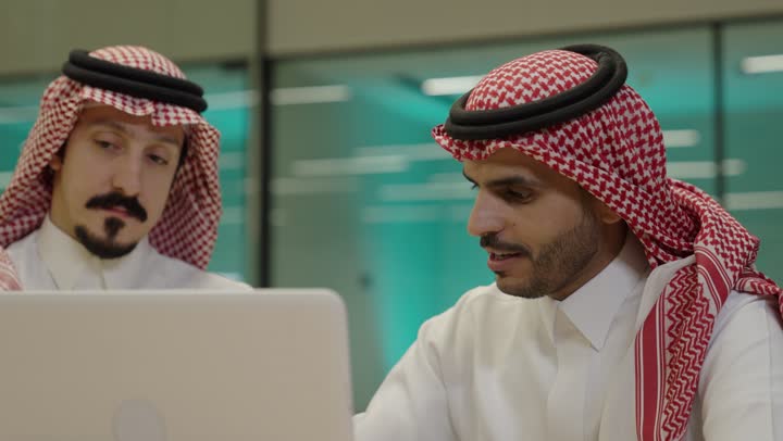 دمج الأعمال مع التقنية و التكنولوجيا، إنجاز الأعمال المطلوبة، رجلان عربيان خليجيان سعوديان يرتديان الزي التقليدي يجلسان في المكتب يعملان على الحاسب المحمول , تحقيق أهداف الشركة, الوحدة والعمل الجماعي، امضاء اوراق العمل