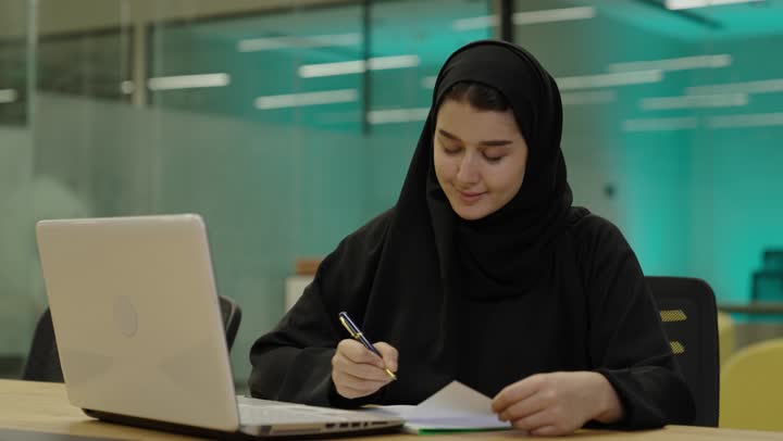 تطور التكنولوجيا وإستخدام الأجهزة التقنية الحديثة،استخدام الورق الأبيض في تسجيل الملاحظات والمعلومات المهمة، مفهوم كتابه وتدوين الملاحظات، امراة عربية خليجية سعودية تجلس على المكتب امام الحاسب المحمول تمسك بيدها قلم تقوم بالكتابة على نوت