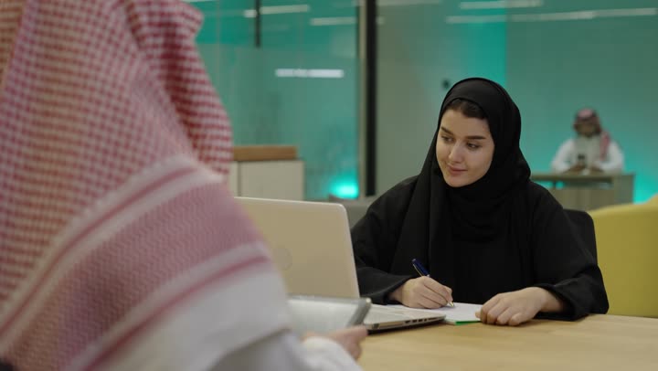 تطور التكنولوجيا وإستخدام الأجهزة التقنية الحديثة, ادخال البيانات من خلال لوحة المفاتيح،  امرأة عربية خليجية سعدية ترتدي العباءة السوداء تقوم بادارة الأعمال من خلال الحاسوب المحمول، كتابة وتدوين الملاحظات، تصوير من الخلف لرجل عربي خليجي سعودي يقوم باستخدام التابلت