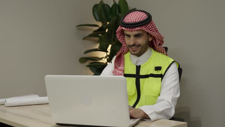 متابعة مخطط المشروع الهندسي اونلاين، مفهوم الهندسة المعمارية، مشاريع إعمارية سعودية وطنية، الاهتمام بالتطور العمراني بسواعد المهندسين السعوديين, مهندس عربي خليجي سعودي يرتدي  الشماغ و سترة الحماية ينجز المهام اليومية عبر الحاسب المحمول