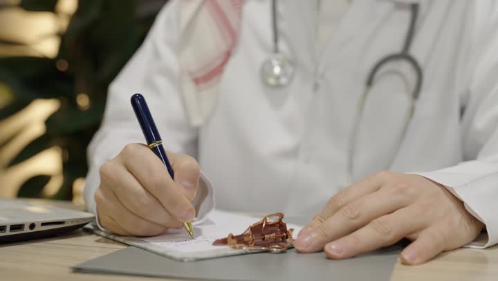 تدوين نتائج الفحوصات بالسجل الطبي، بورتريه مقرب ليدطبيب عربي خليجي سعودي يرتدي المعطف الأبيض يقوم بكتابة و إعداد التقارير الطبية، العمل في القطاع الصحي في المملكة العربيه السعودية