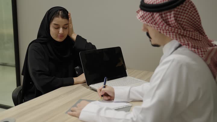 مفهوم التواصل بين الطبيب والمريض، الاستشارة الطبية داخل العيادة، العمل في القطاع الصحي, طبيب عربي خليجي سعودي يرتدي الثوب الابيض و الشماغ و المعطف الطبي يجلس امام الحاسب المحمول يقوم ابلحديث مع المريضة ، اعطاء النصائح و الارشادات، كتابة وتدوين التقارير والحالات الصحية