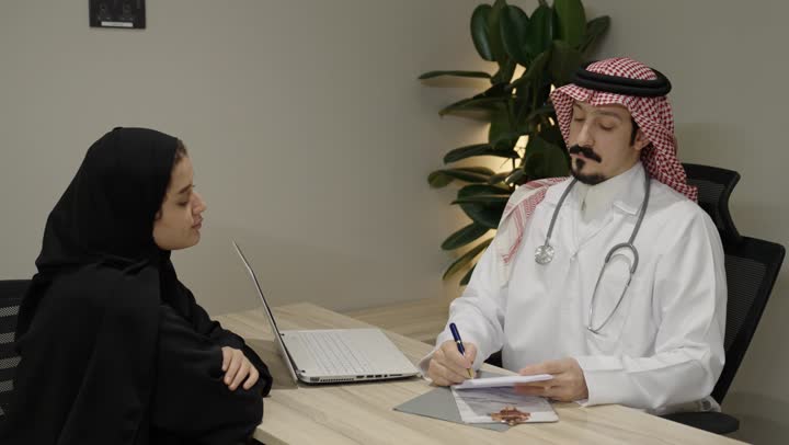 الاستشارة الطبية داخل العيادة، العمل في القطاع الصحي, طبيب عربي خليجي سعودي يرتدي الثوب الابيض و الشماغ و المعطف الطبي يجلس امام الحاسب المحمول يقوم ابلحديث مع المريضة ،  مفهوم التواصل بين الطبيب والمريض، اعطاء النصائح و الارشادات، كتابة وتدوين التقارير والحالات الصحية