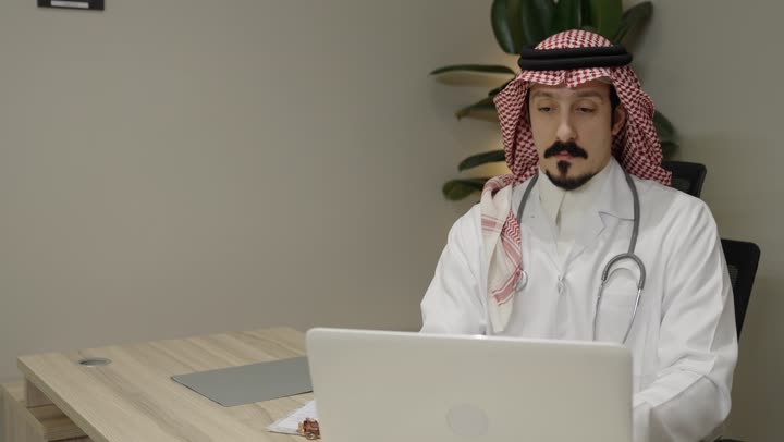 Managing patient files electronically on the laptop, integrating modern technology in the field of medical work, an Arab Gulf Saudi man wearing a ghutrah and a white medical coat with a stethoscope using a laptop, providing therapeutic services in medical centers and hospitals, writing and documenting reports and health cases.