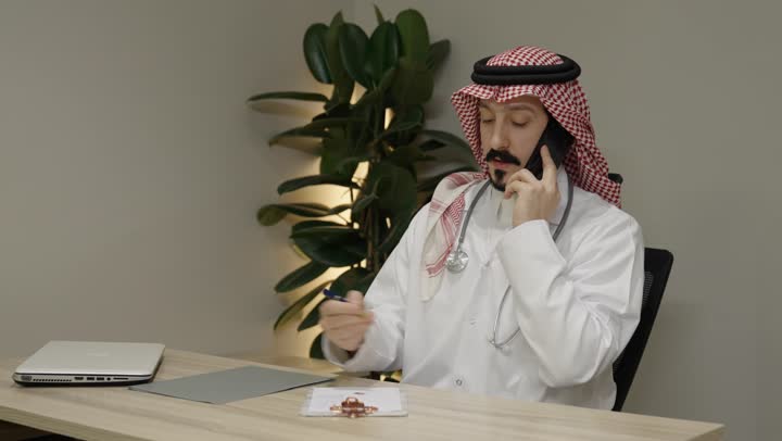 العمل في القطاع الطبي في المملكة العربية السعودية،  كتابة وتدوين التقارير والحالات الصحيه, طبيب عربي خليجي سعودي يرتدي المعطف الطبي و الشماغ يستخدم الهاتف المحمول، اجراء مكالمة هاتفية عبر الجوال