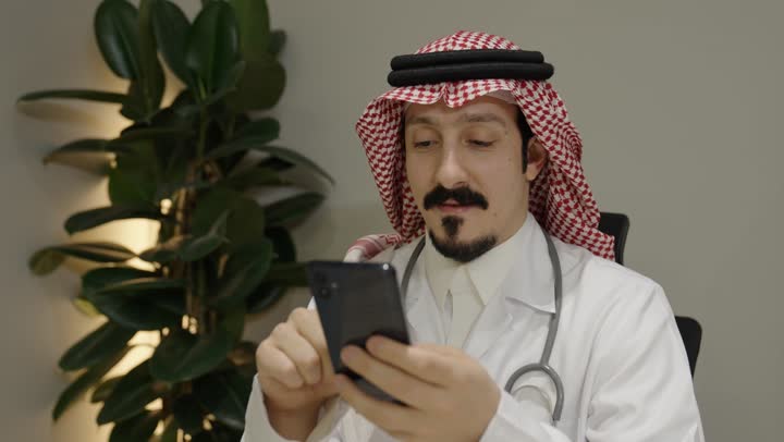 استخدام التقنيات الحديثه في مجال الطب، النقر على شاشة الجوال، تصفح مواقع التواصل الاجتماعي، استخدام جهاز تقني حديث ومتطور، تصوير مقرب لطبيب عربي خليجي سعودي يرتدي الثوب و الشماغ و المعطف الطبي و السماعات يستخدم الهاتف المحمول