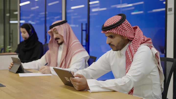 Integrating technology in the development of businesses and companies, professions and administrative jobs in the Kingdom of Saudi Arabia, focus and meticulous work, a group of Arab Gulf employees wearing traditional attire working inside the office, using modern technological devices, writing and taking notes.