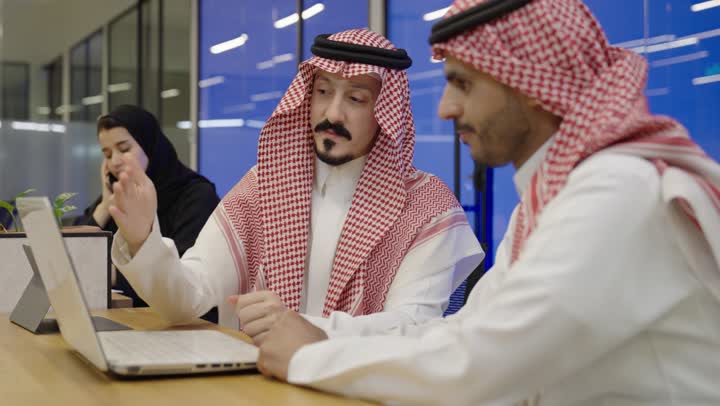 Discussion of plans and explanation of work tasks, business meeting and project completion, use of modern technical devices, exchange of experiences among colleagues, development of businesses and companies, two Arab Gulf Saudi men wearing thobes and ghutras sitting in front of a laptop, an Arab Gulf Saudi woman in a hijab making a phone call via her mobile.