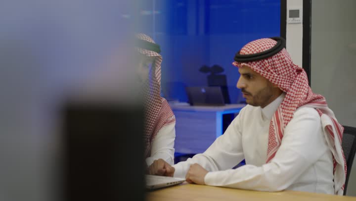 اجتماع عمل وانجاز المشاريع، استخدام الاجهزه التقنية الحديثة، الحوارات والنقاشات ضمن الفريق، تبادل الخبرات بين الزملاء, تطوير الاعمال والشركات, مناقشة الخطط وشرح مهام الاعمال,فريق عربي خليجي سعودي يرتدي الزي التقليدي يجلسون في اجتماع داخل المكتب