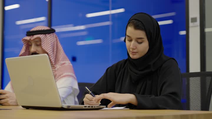 Using a tablet to accomplish work, tapping on the laptop keyboard, using white paper to record notes and important information, the concept of writing and taking notes, an Arab Gulf Saudi woman wearing a hijab and an abaya holding a pen writing on a notebook sitting next to her colleague.