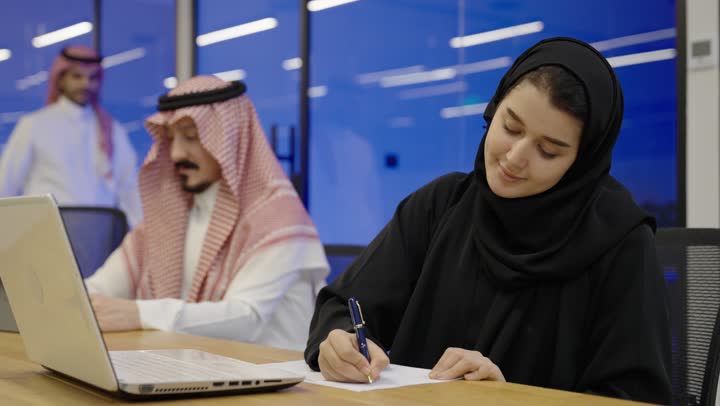 Clicking on the laptop keyboard, using white paper to record notes and important information, the concept of writing and taking notes, an Arab Gulf Saudi woman wearing a hijab and an abaya holding a pen writing on a notebook sitting next to her colleague.