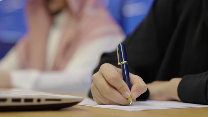 استخدام الورق الأبيض في تسجيل الملاحظات والمعلومات المهمة، مفهوم كتابه وتدوين الملاحظات، تصوير مقرب لامراة عربية خليجية سعودية تمسك بيدها قلم تقوم بالكتابة على نوت