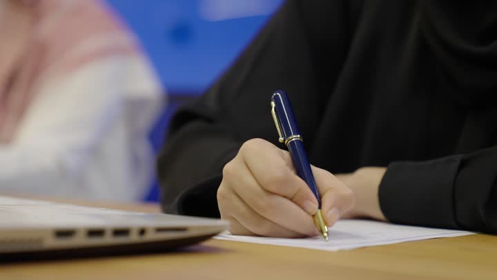 مفهوم كتابه وتدوين الملاحظات،تصوير مقرب لامراة عربية خليجية سعودية تمسك بيدها قلم تقوم بالكتابة على نوت، استخدام الورق الأبيض في تسجيل الملاحظات والمعلومات المهمة