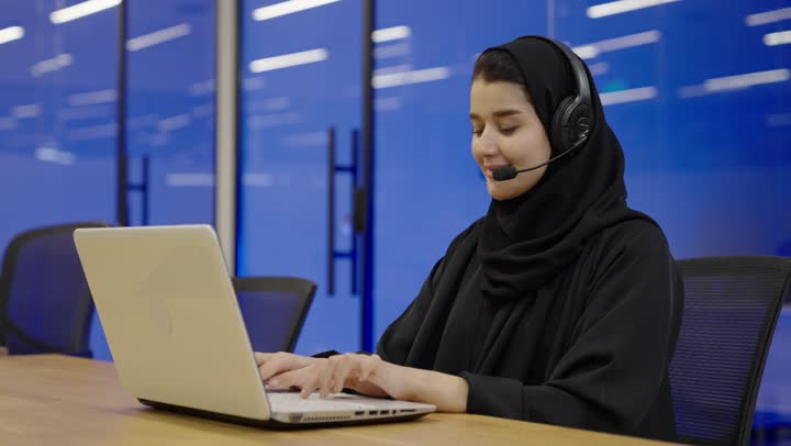 استخدام التقنيات الحديثة والمتطورة، الرد على الاستعلامات والاستفسارات وخدمة العملاء، امراه عربية خليجية سعودية محجبة مبتسمة ترتدي العباءة السوداء تجلس على الكرسي أمام الحاسوب المحمول وتضع سماعة الهيدفون، النقر على لوحة المفاتيح