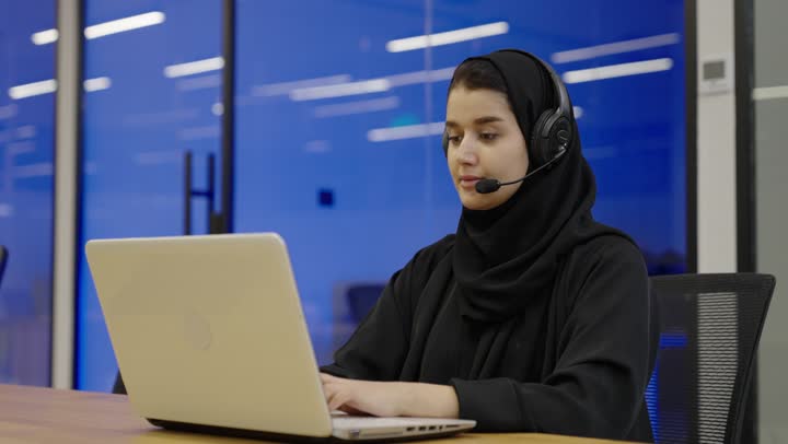 Typing on the keyboard via a laptop, an Arab Gulf Saudi woman wearing a hijab and an abaya, wearing headphones, working in customer service, inquiries and questions, answering customer questions, following up and developing work.