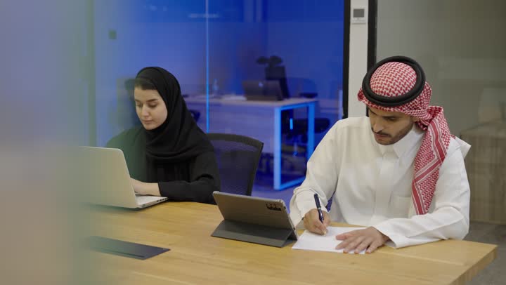 Typing on a keyboard, a Saudi Arabian Gulf woman in a hijab wearing an abaya is using a computer in the office, while a Saudi Arabian Gulf man dressed in a thobe and ghutrah is sitting in front of a tablet writing and taking notes.