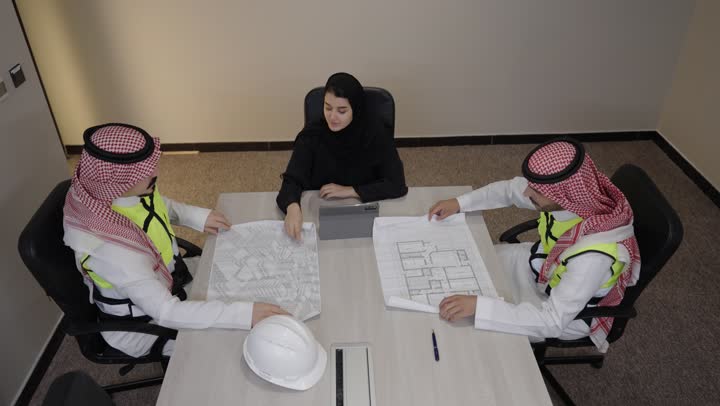 متابعة تنفيذ الخطط الهندسية، الشركات المختصة بورشات البناء والمقاولات، التخطيط ووضع اساسيات المخططات المعمارية، مهندسان عربيان خليجيان سعوديان يرتديان سترة الحماية و الغترة يجلسان داخل غرفه الاجتماع يعملان على مناقشة مخطط الإنشاء وتحديد القياسات بدقة برفقة زميلتهم