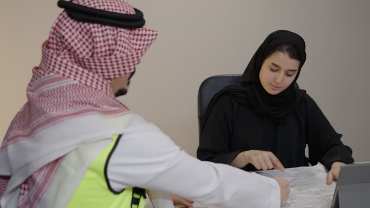 Supervising the project management and follow-up, the concept of engineering and architectural construction, working in the engineering sector, a Saudi Gulf Arab engineer wearing a safety vest and a ghutrah reviewing and presenting project blueprints in full alongside his colleague.