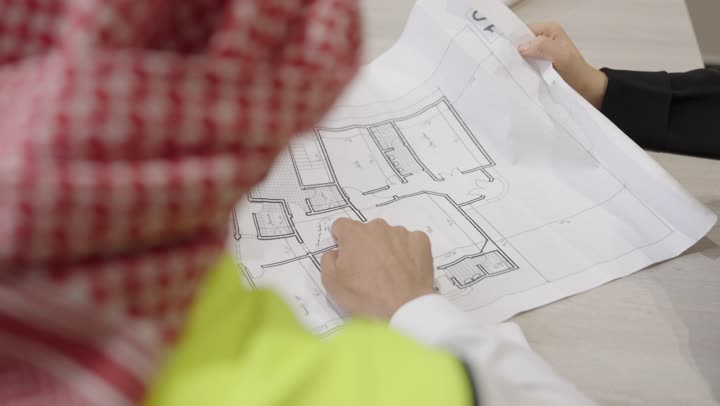 مفهوم الهندسة والانشاءات المعمارية, العمل في القطاع الهندسي, تصوير مقرب لمهندس عربي خليجي سعودي يرتدي ستره الحماية و الغتره يقوم بمراجعة وعرض مخطوطات المشاريع بشكل كامل برفقة زميلته, إدارة ومتابعة المشروع