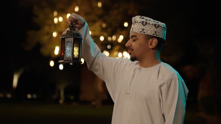 فرحة استقبال شهر رمضان، الأجواء الرمضانية الليلية، تصوير مقرب لرجل عربي خليجي عماني يرتدي الدشداشة و الكمة يمسك بيدية فانوس معدني مزخرف ، النظر للكاميرا بايماءات السعادة و السرور