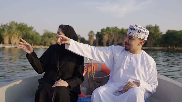 Pointing at something, taking memorable selfies with a mobile phone, documenting moments and building successful social relationships, strengthening marital bonds and ties of love and affection, an Omani Gulf Arab couple wearing traditional dress sitting in a boat, expressions of happiness and joy