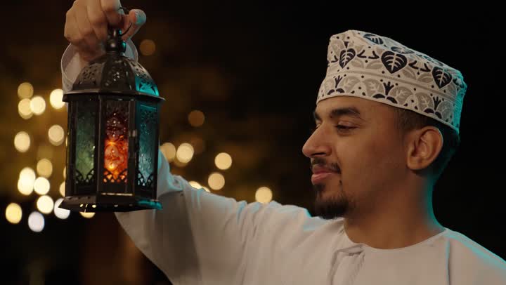 الأجواء الرمضانية الليلية، فرحة استقبال شهر رمضان، تصوير مقرب لرجل عربي خليجي عماني يرتدي الدشداشة و الكمة يمسك بيدية فانوس معدني مزخرف ، النظر للكاميرا بايماءات السعادة و السرور