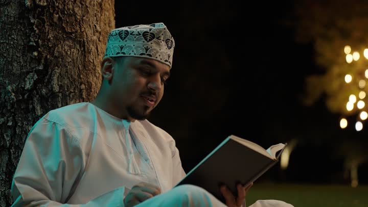 قراءة الكتب بتمعن ، ثقافة التعلم الذاتي، الاستمتاع باوقات الفراغ ، رجل عربي خليجي عماني يرتدي الدشداشة و الكمة يجلس فى الهواء الطلق يحمل بيده كتاب تثقيفي، ايماءات التركيز والاندماج ، قراءة الروايات والقصص