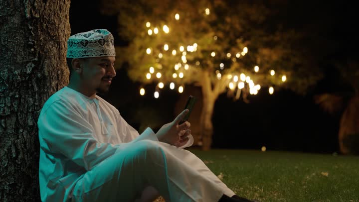 العمل عن بعد، التواصل وتبادل الأحاديث والأخبار مع الأهل والأصدقاء، رجل عربي خليجي عماني يرتدي الدشداشة و الكمة يجلس فى الهواء الطلق يمسك بيده الهاتف المحمول، استخدام جهاز تقني حديث ومتطور، تصفح التطبيقات وبرامج التواصل الاجتماعي