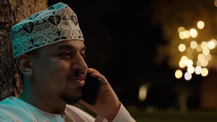 إجراء مكالمه صوتية عبر الهاتف المحمول، قضاء وقت ممتع في الهواء الطلق، استخدام الاجهزة التقنية الحديثة، تصويرمقرب لرجل عربي خليجي عماني الدشداشة و الكمة يمسك بيدية الهاتف المحمول، دردشه ومحادثات مع الاصدقاء والاقارب