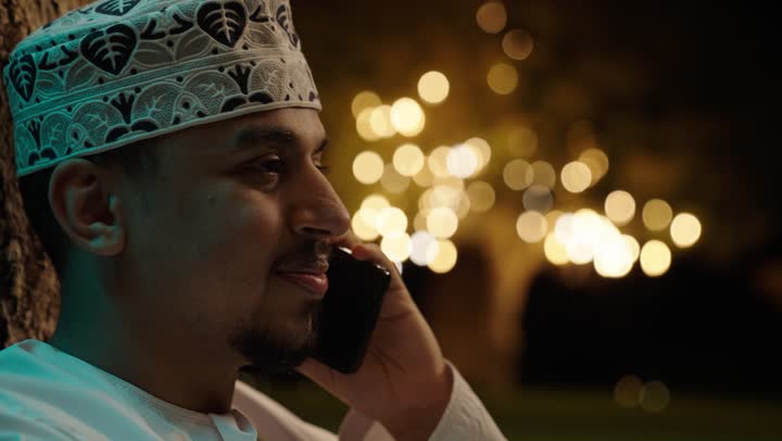 Enjoying time outdoors, using modern technology, making a voice call on a mobile phone, a close-up photograph of an Omani Gulf Arab man in a dishdasha and kuma holding a mobile phone, chatting and having conversations with friends and relatives.
