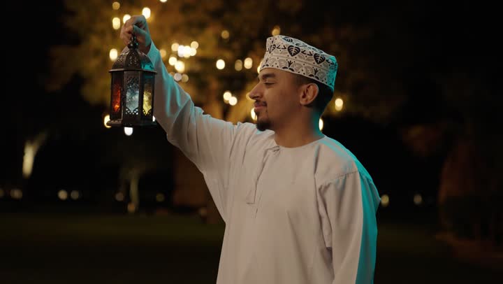 النظر للكاميرا بتعابير السعادة و السرور، البهجة بقدوم شهر الخير، التحضير لاستقبال رمضان المبارك, رجل عربي خليجي عماني يرتدي الدشداشة و الكمة يمسك فانوس معدني مزخرف و ينظر الية, الاحتفال بالمناسبات الدينية الإسلامية