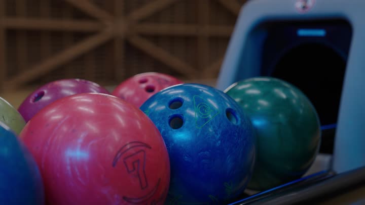 رجل عربي سعودي خليجيى يمسك كرة البولينغ المعدنية الزرقاء ,مجموعة كرات منظمة و مرتبة استعدادًا لانطلاق المباراة , نشاط عائلي ترفيهي في صالة البولينج