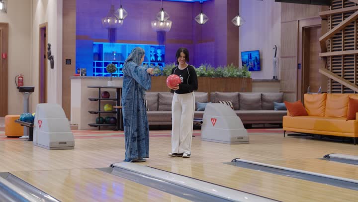 امرأة عربية خليجية سعودية محجبة ترتدي عباءة زرقاء تشرح لابنتها الطريقة الصحيحة لرمي كرة البولينغ ,ابنة ترتدي الزى الكاجوال ترمى الكرة باحترافية ,التصفيق و التهليل بعد تحقيق الفوز ,مفهوم الاسرة السعيدة , نشاط ترفيهي عائلي في صالة البولينج