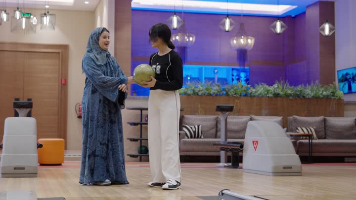 فتاة عربية خليجية سعودية ترتدي الزي الكاجوال ترمي كرة البولينغ المعدنية تقف عند نقطة البداية ,ام عربية محجبة ترتدي عباءة زرقاء تشاهد ابنتها اثناء اللعب ,الفرحة و السعادة عند تحقيق الفوز ,مفهوم الاسرة السعيدة , نشاط ترفيهي عائلي في صالة البولينج