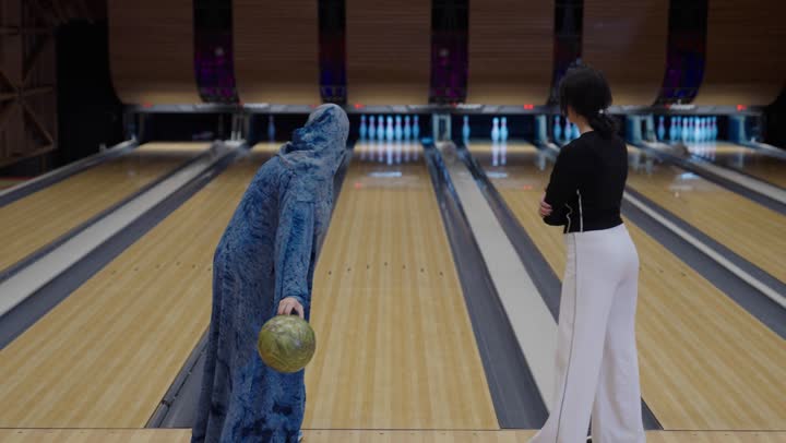 فتاة عربية خليجية سعودية ترتدي الزي الكاجوال تشاهد والدتها اثناء لعب رياضة البولينغ ,امرأة عربية محجبة ترتدي العباءة الزرقاء ترمي الكرة المعدنية لاسقاط الدبابيس البلاستيكية ,التصفيق و السعادة بعد تحقيق الفوز, رفع اصبع الابهام في اشارة لاجادة طريقة اللعب , نشاط ترفيهي عائلي في صالة البولينج