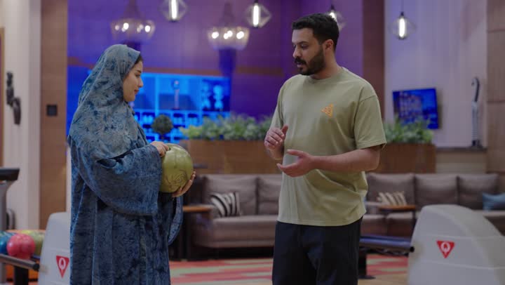 زوجان عربيان يقضيان اوقات عائلية مسلية اثناء ممارسة رياضة البولينغ ,زوج عربي خليجي سعودي يقوم بشرح القواعد الاساسية لللعب ,زوجة عربية خليجية سعودية محجبة ترتدي عباءة باللون الازرق تستمع لزوجها بابتسامة في جو اسري جميل , نشاط ترفيهي عائلي في صالة البولينج