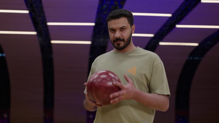 رجل عربي خليجى سعودي يرتدي الزي الكاجوال يمسك كرة البولينغ باحترافية واتقان ,لاعب محترق يحرك الكرة بين يديه ,نظرة اصرار وعزيمة على تحقيق النتيجة الكاملة , نشاط ترفيهي عائلي في صالة البولينج