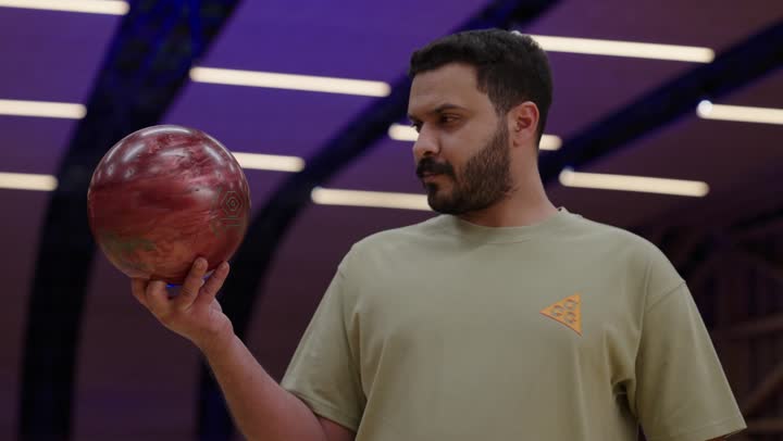 النظر لكرة البولينع بروح العزيمة و التصميم على الفوز ,رجل عربي سعودي خليجي يمسك كرة البولينغ الحمراء مع نظرة حادة مصرة على تحقيق الانتصار , نشاط ترفيهي عائلي في صالة البولينج
