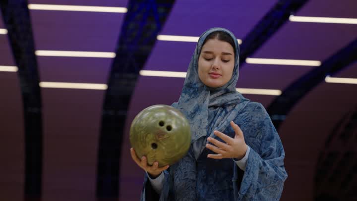 طريقة احترافية متقنة أثناء ممارسة الرياضة , امرأة عربية خليجية سعودية محجبة ترتدي عباءة باللون الازرق تلعب بكرة البولينغ بين يديها ,قضاء أوقات تنافسية مسلية مع العائلة , نشاط ترفيهي عائلي في صالة البولينج