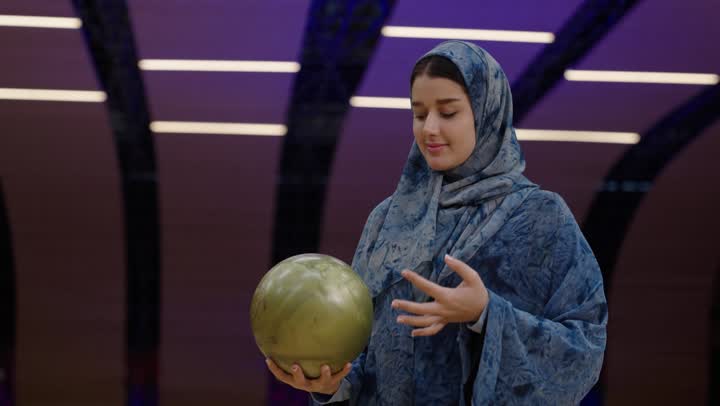 الاتقان و الاحترافية أثناء ممارسة رياضة البولينغ , امرأة عربية خليجية سعودية محجبة ترتدي عباءة باللون الازرق تحمل كرة صفراء بين يديها ,التحدي و الاصرار على الفوز , نشاط ترفيهي عائلي في صالة البولينج