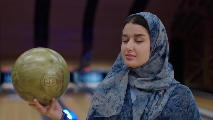 سيدة عربية خليجية سعودية محجبة ترتدي عباءة زرقاء اللون تحمل كرة بولينغ صفراء اللون ,نظرة التحدي و الاصرار على الفوز ,مفهوم المنافسة مع قضاء أوقات ممتعة مع العائلة , نشاط ترفيهي عائلي في صالة البولينج