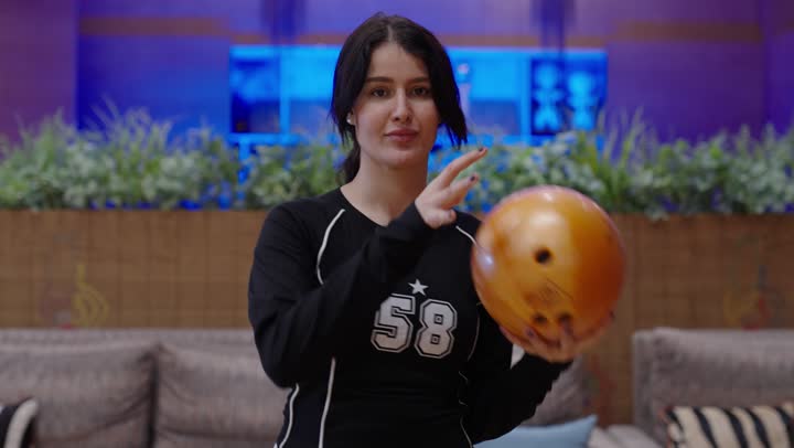 مفهوم الثقة بالنفس والعزيمة على الفوز ,فتاة عربية خليجية سعودية ترتدي الزي الكاجوال تحمل كرة بولينغ صفراء باتقان ,اضاءة زرقاء في غرفة المعيشة ,ابتسامة الثبات والسعادة , نشاط ترفيهي للعائلة في صالة البولينج