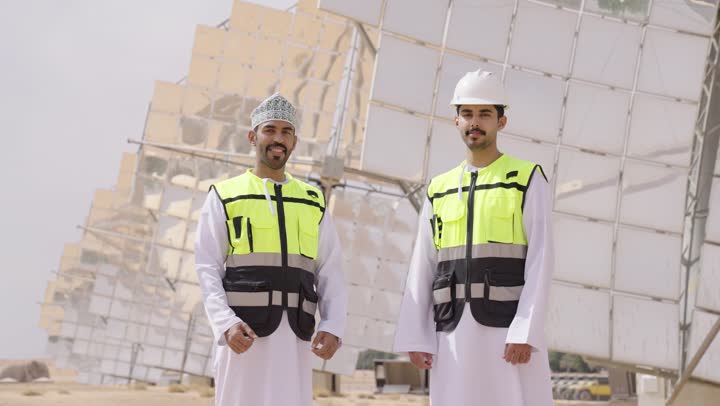 مهندسان عربيان خليجيان عمانيان يشرفان على محطات الطاقة الشمسية يقفان بأيدى متقاطعة, إرتداء خوذة وسترة الحماية الخاصة بالعمل، مخططات وتصاميم هندسية، حوارات ونقاشات عمل ، محطات الطاقة الشمسية المتجددة، الواح وستلايت انتاج الطاقة الكهربائية