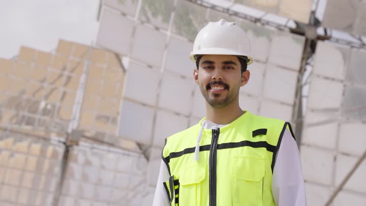 الوقوف بأيدى متقاطعة, الإشراف على محطات توليد الكهرباء, استخدام التقنية والتكنولوجيا في المشاريع المستقبلية,  مهندس طاقة عربي خليجي عمانى يحمل بيده جهاز تقني ينظر الى الكاميرا، إرتداء خوذة وسترة الحماية الخاصة بالعمل