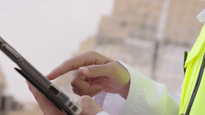 تصوير مقرب ليد مهندس طاقة عربي خليجي عمانى يحمل بيده جهاز تقني، إرتداء خوذة وسترة الحماية الخاصة بالعمل, الإشراف على محطات توليد الكهرباء, استخدام التقنية والتكنولوجيا في المشاريع المستقبلية, التدقيق والإشراف على العمل