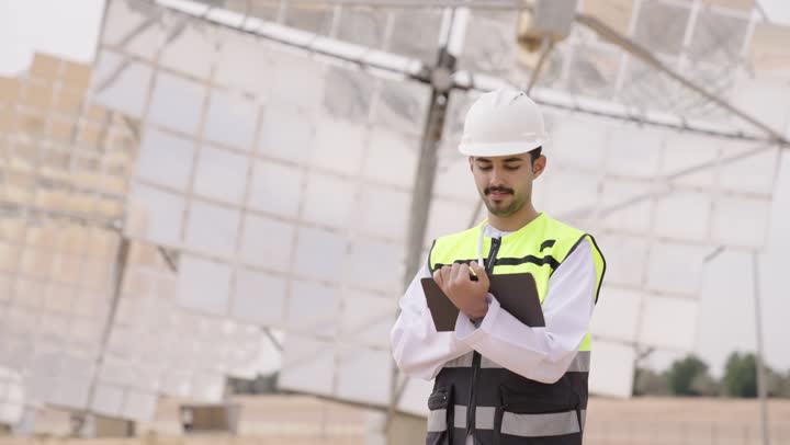 Auditing and supervising work, solar power stations, auditing observations, an Arab Gulf Omani energy engineer holding a technical device in his hand, wearing a helmet and work safety vest.