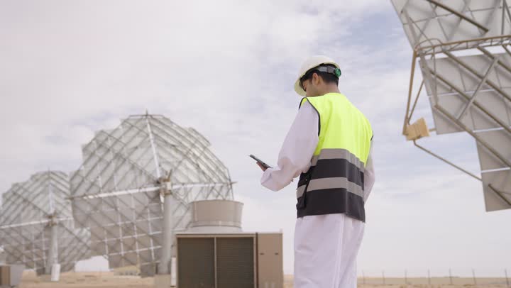 دمج التقنيات الحديثة و المتطورة بالأعمال الهندسية, تصوير من الخلف لمهندس طاقة عربي خليجي عمانى يحمل التابلت، إرتداء خوذة وسترة الحماية الخاصة بالعمل