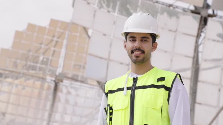Looking at the camera with confident expressions, supervising power generation stations, using technology and tech in future projects, an Arab Gulf Omani energy engineer standing with crossed arms, wearing a helmet and work safety vest.
