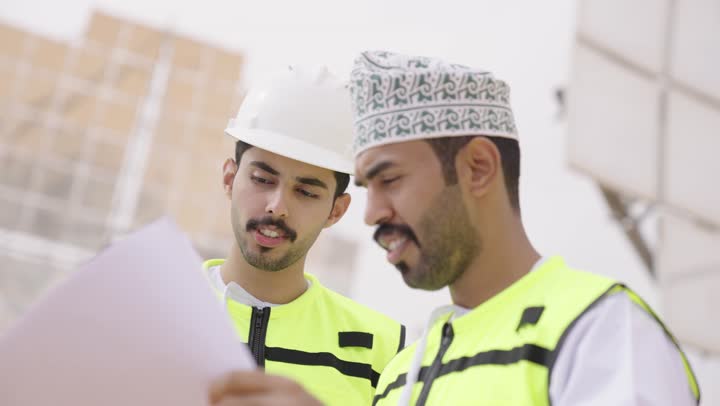 الإشراف على العمل، ستلايت، أنظمة الطاقة الشمسية,  تبادل الخبرة و الرأى بين الزملاء, مهندسان عربيان خليجيان عمانيان يشرفان على محطات الطاقة الشمسية يحملان مخطط و تصميم هندسى، إرتداء خوذة وسترة الحماية